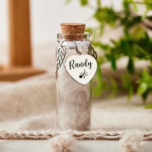 K&ouml;nnte beinhalten: Ein kleines Glasgef&auml;&szlig; mit einem Korkverschluss, gef&uuml;llt mit wei&szlig;em Fell. Das Gef&auml;&szlig; hat einen silbernen Herzanh&auml;nger mit dem Namen "Randy" und einem Sternschnuppen-Design. Der Anh&auml;nger ist an einem silbernen Engelsfl&uuml;gel-Anh&auml;nger befestigt.