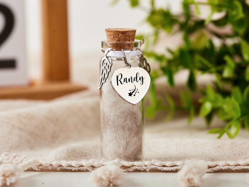 K&ouml;nnte beinhalten: Ein kleines Glasgef&auml;&szlig; mit einem Korkverschluss, gef&uuml;llt mit wei&szlig;em Fell. Das Gef&auml;&szlig; hat einen silbernen Herzanh&auml;nger mit dem Namen "Randy" und einem Sternschnuppen-Design. Der Anh&auml;nger ist an einem silbernen Engelsfl&uuml;gel-Anh&auml;nger befestigt.