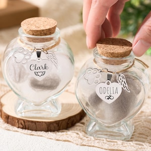 May include: Two heart-shaped glass jars with cork stoppers, filled with a light-colored substance. Each jar has a silver charm with a name, paw print, and wing design. One jar is on a wooden slice.