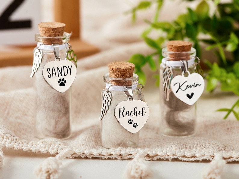 K&ouml;nnte beinhalten: Drei kleine Glasflaschen mit Korkstopfen und wei&szlig;em Band. Jede Flasche enth&auml;lt eine kleine Menge wei&szlig;en Fells und hat einen silbernen Herzanh&auml;nger mit einem eingravierten Namen. Die Namen sind Sandy, Kevin und Rachel. Jeder Anh&auml;nger hat auch einen kleinen Pfotenabdruck.