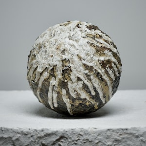 Op de afbeelding: Een bolvormig, getextureerd object met een donkerbruine basis en witte, druppelachtige patronen. Het rust op een vierkant, getextureerd, wit blok tegen een grijze achtergrond. De uitstraling is minimalistisch en artistiek.