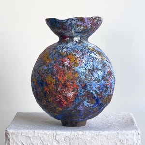 Puede incluir: Un jarrón esférico de cerámica con superficie texturizada. El jarrón presenta una abertura ancha y acampanada y está pintado con un diseño abstracto colorido en tonos azules, rojos, naranjas y morados. El jarrón se exhibe sobre un pedestal blanco.