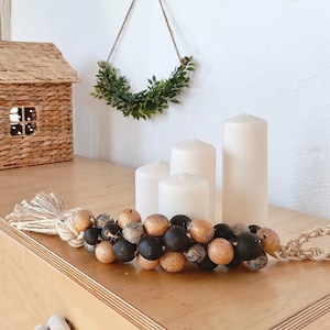 Może przedstawiać: Girlanda z drewnianych koralików z czarnymi i złotymi koralikami, frędzlem i domem w stylu wiejskim w tle. Trzy białe świece stoją na drewnianej powierzchni.