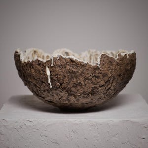 Op de afbeelding: Een kom met een ruwe, donkerbruine, getextureerde buitenkant en een contrasterende gladde, witte binnenkant. De rand is ongelijk en grillig, met druppels witte glazuur die langs de donkere, gekraterde buitenwand van de kom naar beneden lopen.