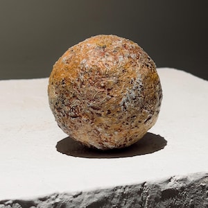 May include: A spherical, textured object with a mottled appearance. The colors range from tan and brown to hints of red and white. The object is resting on a white surface, with a dark background.