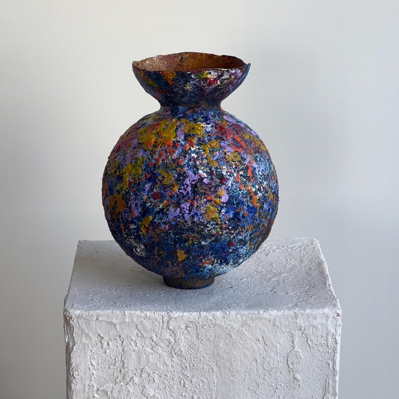K&ouml;nnte beinhalten: Eine handgefertigte, kugelf&ouml;rmige Vase mit einer strukturierten, mehrfarbigen Oberfl&auml;che. Die Vase hat einen dunkelblauen Sockel, verziert mit Spritzern in Rot, Gelb und Lila. Die obere &Ouml;ffnung hat eine kontrastierende braune Farbe. Die Vase steht auf einem wei&szlig;en, strukturierten Sockel.