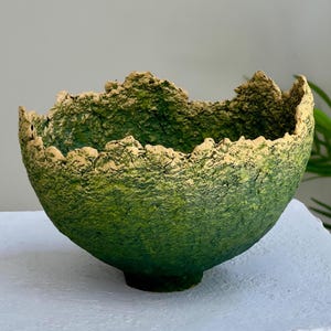 Puede incluir: Un cuenco verde texturizado con un borde áspero e irregular. El cuenco tiene forma redondeada y una base verde oscuro. El borde es de un color verde amarillento más claro. El cuenco parece estar hecho de un material natural.