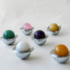 May include: A collection of decorative objects resembling miniature planets. Each planet features a polished, colorful gemstone sphere atop a silver-colored metal base. The gemstones are various colors, including green, pink, purple, yellow, white, and orange.