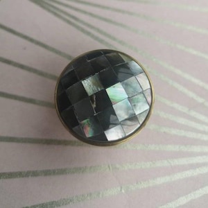 Puede incluir: Perilla redonda y decorativa con una superficie de mosaico de cuadrados grises oscuros e iridiscentes. La perilla tiene un borde color latón y probablemente sea para muebles o armarios.