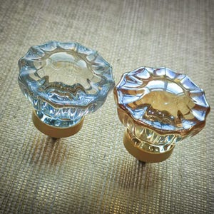 May include: Two decorative glass cabinet knobs. One is clear with a blue tint, the other is amber. Both have a scalloped edge and sit on a gold-colored base. The knobs are displayed on a textured surface.