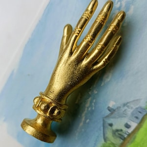 May include: A gold-colored hand sculpture with detailed finger and wrist accents. The hand is posed in an open gesture, with a textured surface. The sculpture is set against a backdrop of a watercolor painting with blue and green hues.