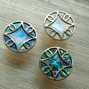 May include: Three decorative cabinet knobs with a round brass frame and iridescent abalone shell inlay. The knobs feature a central diamond shape surrounded by a decorative openwork design. The abalone shell displays shades of blue, green, and white.
