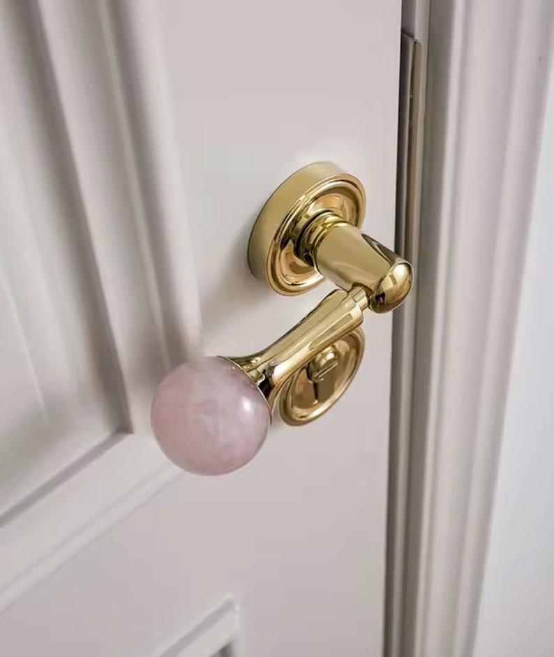 May include: A gold door handle with a pink, round knob. The handle is attached to a white door.
