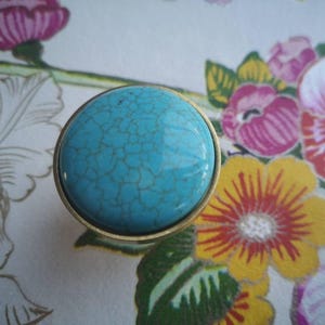 May include: A round cabinet knob with a turquoise stone center and a gold-colored metal rim. The knob has a cracked, textured appearance. The background features a floral pattern with pink, yellow, and green flowers.