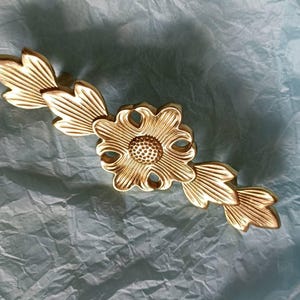 May include: A gold-coloured decorative floral accent featuring a central flower and leaf-like extensions. The flower has a detailed centre and petal design. The piece is likely made of metal against a blue background.