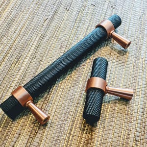 May include: Two pieces of furniture hardware: a long handle and a smaller knob. Both have a black, textured cylindrical body and copper-coloured accents. The handle is approximately 20 cm long, while the knob is about 8 cm. Suitable for cabinets or drawers.