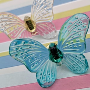 Puede incluir: Dos adornos decorativos de mariposa. Una mariposa tiene alas azules con detalles verdes, la otra tiene alas rosas y amarillas. Ambas tienen detalles centrales dorados y verdes. Las mariposas están sobre un fondo a rayas.