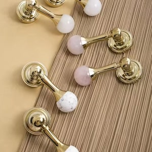 May include: Six gold cabinet knobs with different coloured stone accents. The knobs have a round base and a long, thin stem. The stone accents are white, pink, and a mix of white and grey.