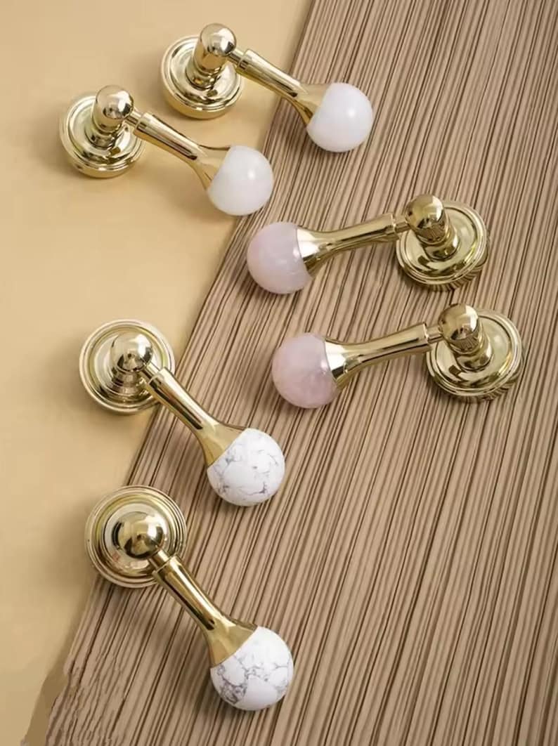 May include: Six gold cabinet knobs with different colored stone accents. The knobs have a round base and a long, thin stem. The stone accents are white, pink, and a mix of white and gray.