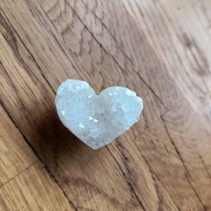 May include: A heart-shaped crystal cluster in a pale blue shade. The crystal has a rough, textured surface with numerous small, sparkling facets. The heart rests on a wooden surface with a warm, brown tone.
