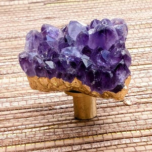 Puede incluir: Pomo decorativo para armario con un grupo de cristales de amatista de color púrpura intenso, con base y tallo dorados. La amatista es de un color rico y vibrante, y el pomo se encuentra sobre un fondo natural.
