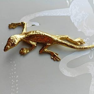 Puede incluir: Figura de lagarto de metal dorado. El lagarto está en posición de reptar, con las patas extendidas y la cola curvada. La superficie del lagarto tiene un aspecto texturizado. La figura está sobre un fondo gris claro.