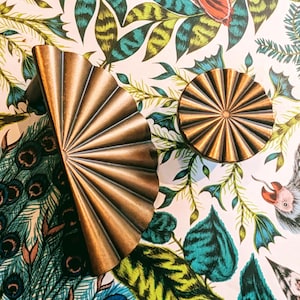 Puede incluir: Dos tiradores de armario en forma de abanico de color bronce y un pomo redondo se muestran sobre un fondo vibrante y estampado. El fondo presenta una mezcla de plumas de pavo real, hojas e ilustraciones de animales en tonos azules, verdes y naranjas.
