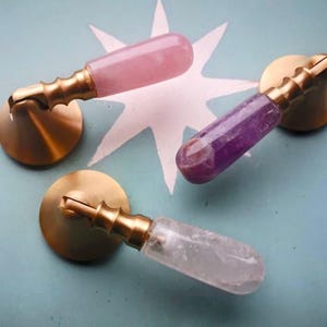 May include: Three decorative cabinet pulls with brass fittings and gemstone handles. One has a pink handle, one a purple handle, and one a clear handle. They are set against a light blue background with a white star design.