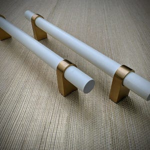 May include: Two cabinet pulls with a white cylindrical handle and gold-colored brackets. The handles are horizontal, and the brackets are rectangular. The pulls are set against a neutral background.