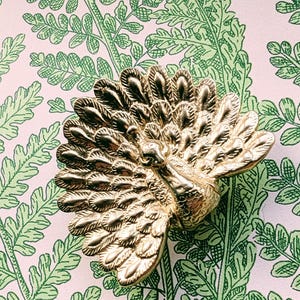 May include: A gold-coloured peacock-shaped cabinet knob. The knob has a detailed fanned tail and is set against a pink background with a green fern pattern.