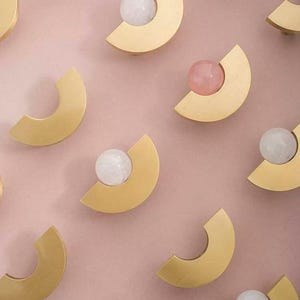 Puede incluir: Herrajes decorativos para armarios con un diseño semicircular dorado y un acento esférico blanco o rosa. Los herrajes están dispuestos sobre un fondo rosa claro, creando una estética moderna y elegante.