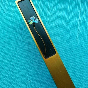 May include: A rectangular letter opener with a gold-colored handle and a black inlay. The inlay features a floral design with iridescent petals and a thin, curved stem. The opener is set against a turquoise background.