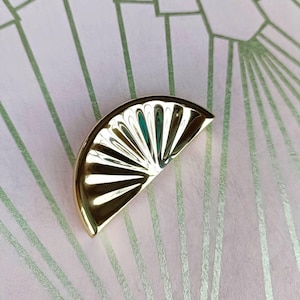 May include: A gold-coloured, fan-shaped decorative object with a reflective surface. The object is set against a pink background with a green art deco pattern. The fan shape has a series of vertical lines.