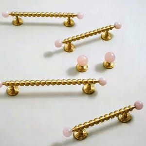 May include: Gold-coloured cabinet hardware featuring a beaded design and pink gemstone accents. The set includes bar pulls and round knobs, perfect for adding a touch of elegance to furniture or cabinetry.