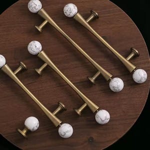 May include: Set of six cabinet pulls with a gold finish and white marble-look spheres. The pulls are arranged on a dark brown wooden surface, showcasing their design and potential use in home decor.