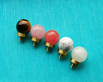 May include: Five decorative knobs with brass bases and gemstone spheres in various colors: tiger's eye, pink, red, white with gray marbling, and clear white. The knobs are arranged diagonally on a turquoise fabric.