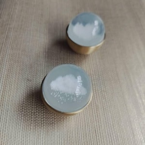 Puede incluir: Dos pendientes redondos con temática de nubes. Cada pendiente presenta una parte superior transparente en forma de cúpula con un diseño de nube blanca y una base dorada. Los pendientes están sobre un fondo neutro.