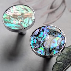 Peut inclure: Deux boutons de meuble ronds avec des incrustations de coquillages d'ormeau irisés. Les boutons ont un bord et une tige en métal argenté. La coquille d'ormeau affiche un mélange de teintes bleues, vertes et violettes. Sur fond floral.