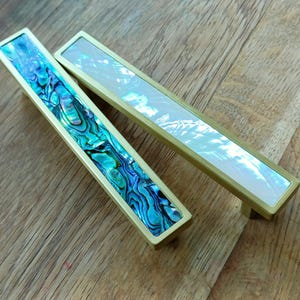 May include: Two rectangular cabinet pulls with brass frames. One has a vibrant blue and green abalone shell inlay, the other a pearlescent white inlay. The pulls are set against a wooden surface.