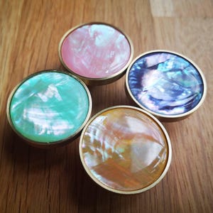 May include: Four round cabinet knobs with iridescent, pearlescent centres in pink, teal, blue, and orange, set in brass-coloured metal. The knobs are arranged on a wooden surface, showcasing their colourful, decorative design.