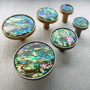 May include: A selection of cabinet knobs with iridescent abalone shell inlays. The knobs have varying sizes and metal finishes, including gold and silver. The abalone shells display a mix of green, blue, and pink hues.