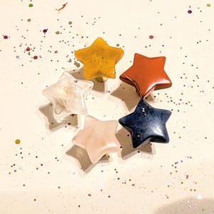 May include: A collection of six star-shaped gemstones in a circular arrangement. The stars are in various colors, including yellow, red, blue, pink, and clear. The background is white with scattered glitter.