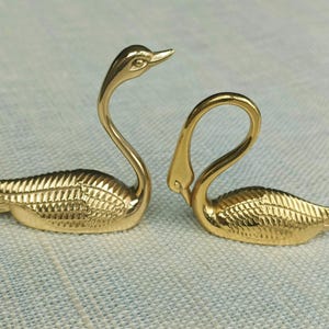 May include: Two gold-colored swan figurines. The swans have long, curved necks and detailed feather patterns on their bodies. One swan has its head up, while the other has its head bent down, creating a graceful pose. Decorative items.