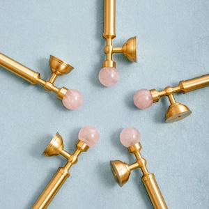 May include: Five gold-coloured metal facial rollers, each with a pink gemstone sphere. The rollers are arranged in a star shape against a light blue backdrop. Each roller is approximately 15 cm in length.