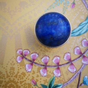 May include: A close-up of a round, deep blue stone with a silver rim. The stone is set against a gold background with floral and leaf patterns in pink, green, and blue.