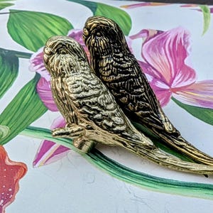 May include: Two decorative bird ornaments, one gold and one bronze, perched on a branch. The birds have textured feather detailing. The background features a floral design with pink and green elements.