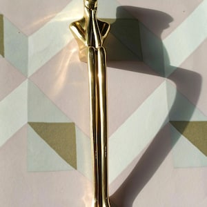 May include: A gold-colored decorative object in the shape of a stylized human figure. The figure has a bust and long, slender legs. The arms are raised to the face. The object is made of a shiny, reflective material.