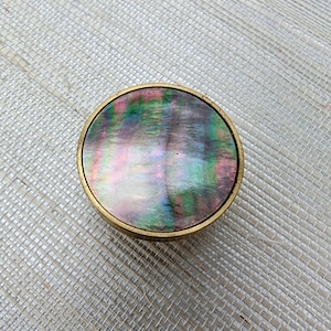 Puede incluir: Una caja decorativa redonda con un borde de metal dorado. La parte superior de la caja presenta una superficie brillante e iridiscente con tonos rosas, verdes y morados. La caja probablemente se utiliza para guardar objetos pequeños.