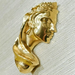 May include: A gold-coloured brooch depicting a classical female bust. The brooch shows detailed facial features, including a profile of the face, hair, and a draped neck. The metal has a polished, reflective surface.