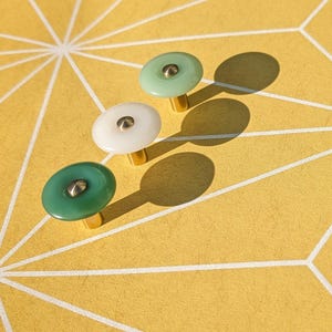 May include: Three decorative cabinet knobs in shades of green and white with gold-colored bases. The knobs are arranged on a yellow surface with a geometric white pattern. The knobs cast long shadows.